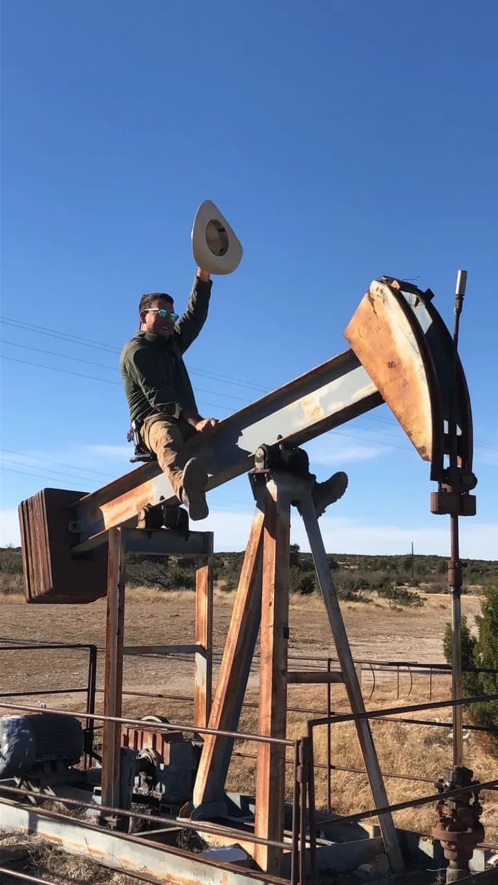 Michael McCammon sitting on the beam of a working pumpjack in West Texas brush country, raising his hat.
